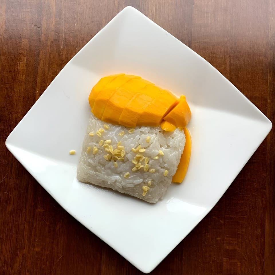 Ddee Thai's mango sticky rice dessert with sliced mango and toasted mung beans on a white plate.
