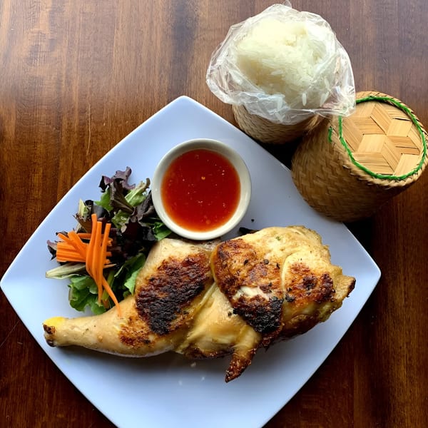 Grilled chicken with sweet chili sauce, salad, and sticky rice in bamboo baskets on a wooden table.